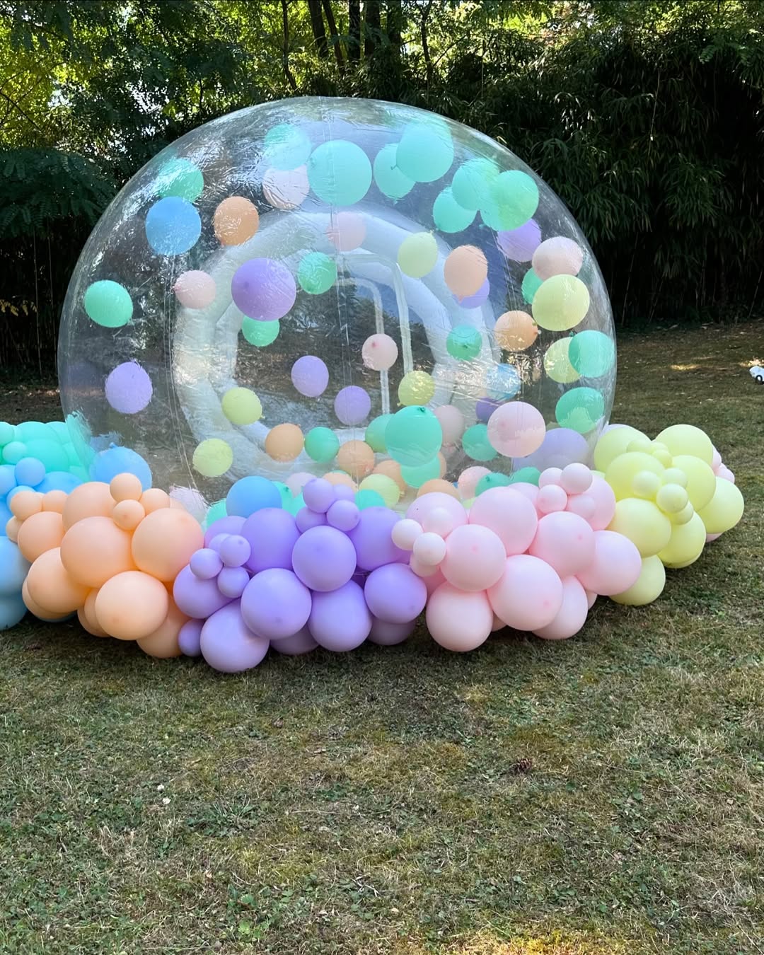 Grande bulle gonflable transparente remplie de ballons pastel, entourée d'une arche de ballons multicolores sur une pelouse verte.