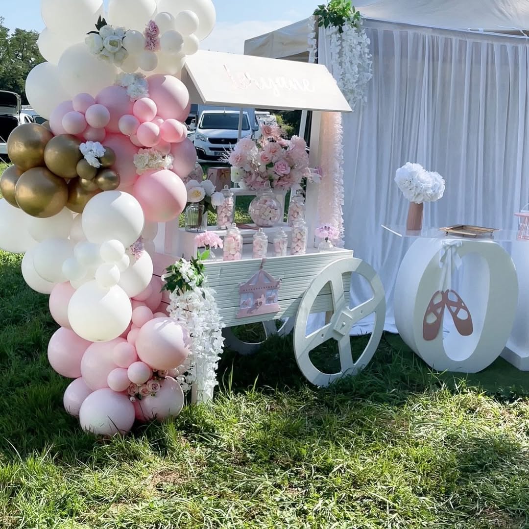 Décoration de fête extérieure thème ballerine avec guirlande ballons rose, blanc, or et un chariot à bonbons, près d'un fond de drapé blanc.