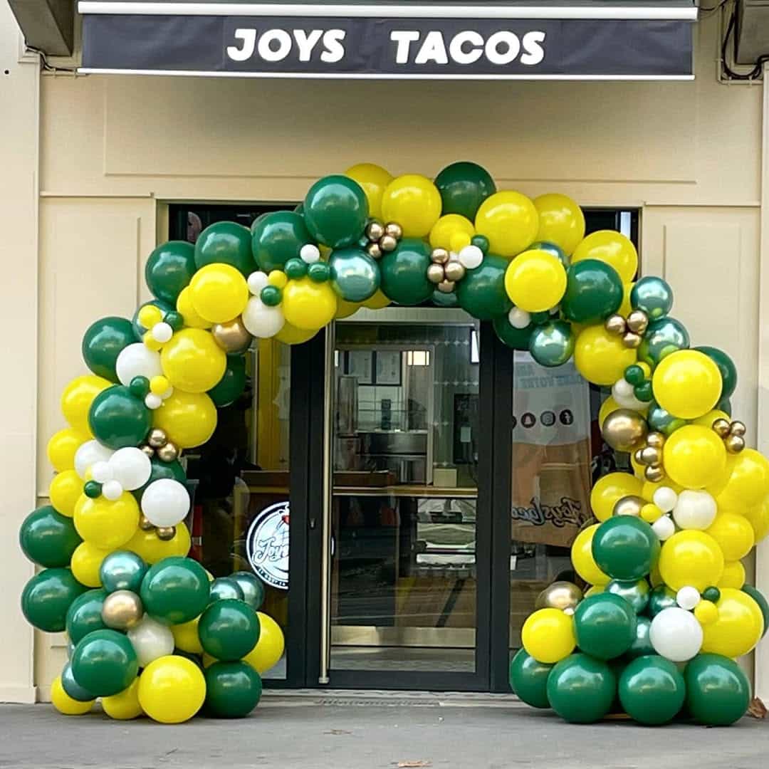 Arche de ballons jaune, vert et or pour l'inauguration de Joys Tacos, décorant l'entrée d'un magasin.