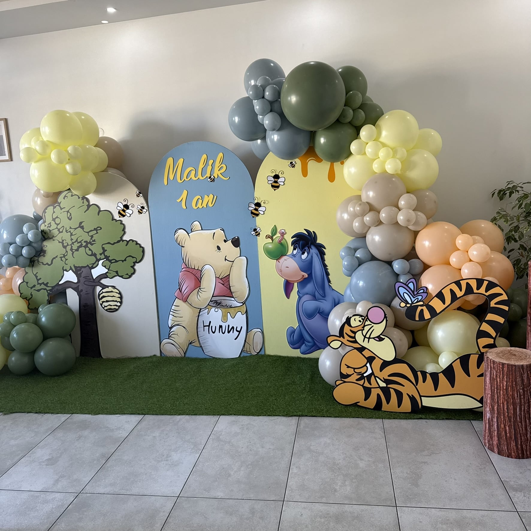 Décoration d'anniversaire Winnie l'Ourson "Malik 1 an" avec Pooh, Eeyore et Tigrou. Arches de ballons vert olive, bleus et jaunes sur gazon artificiel.