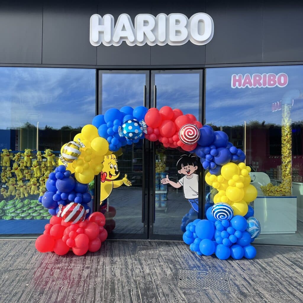 Magasin Haribo : L'entrée festive avec Goldbear et ballons. Entrée du magasin HARIBO sous le ciel bleu. Grande arche de ballons rouge, jaune et bleu, avec Goldbear et un garçon accueillant les clients.