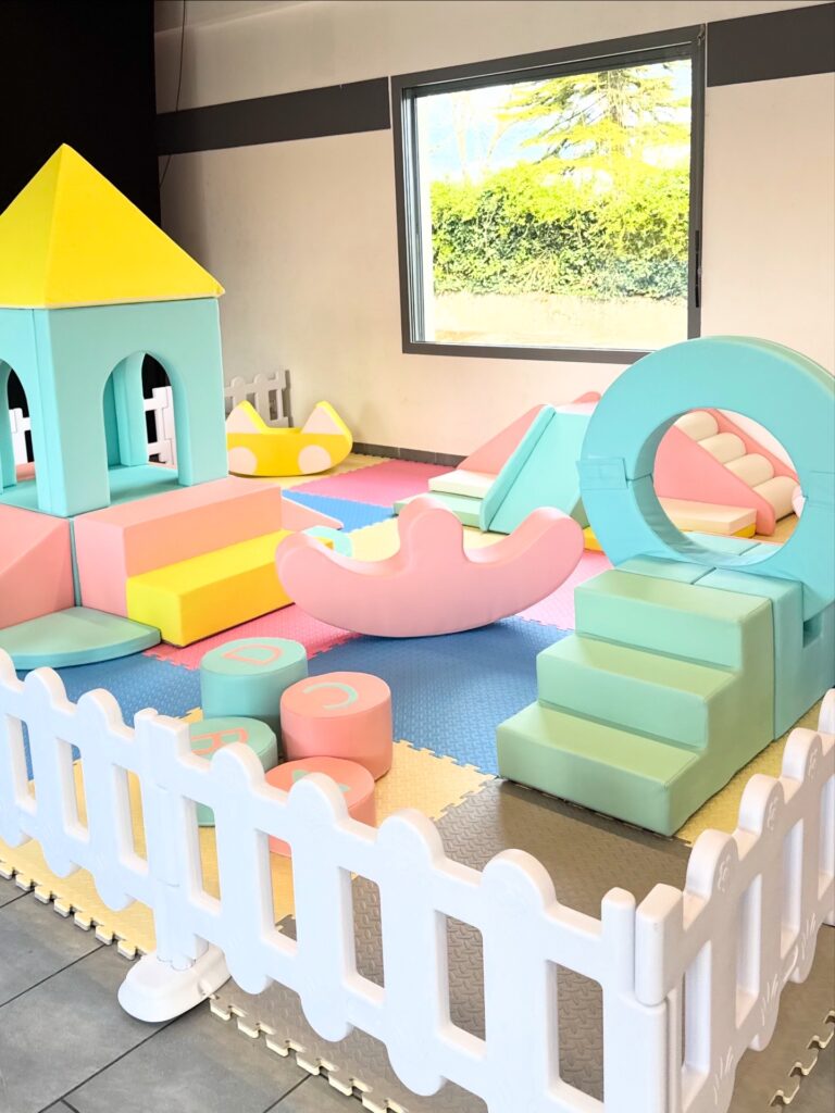 Aire de Jeux Intérieure en Mousse Pastel pour Enfants Aire de jeux intérieure en mousse pastel pour enfants avec château, toboggan et clôture blanche sécurisée.